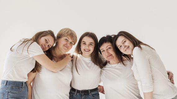 Fünf Frauen unterschiedlicher Altersgruppen umarmen sich vor weißem Hintergrund. Sie tragen weiße T-Shirts und Jeans.
