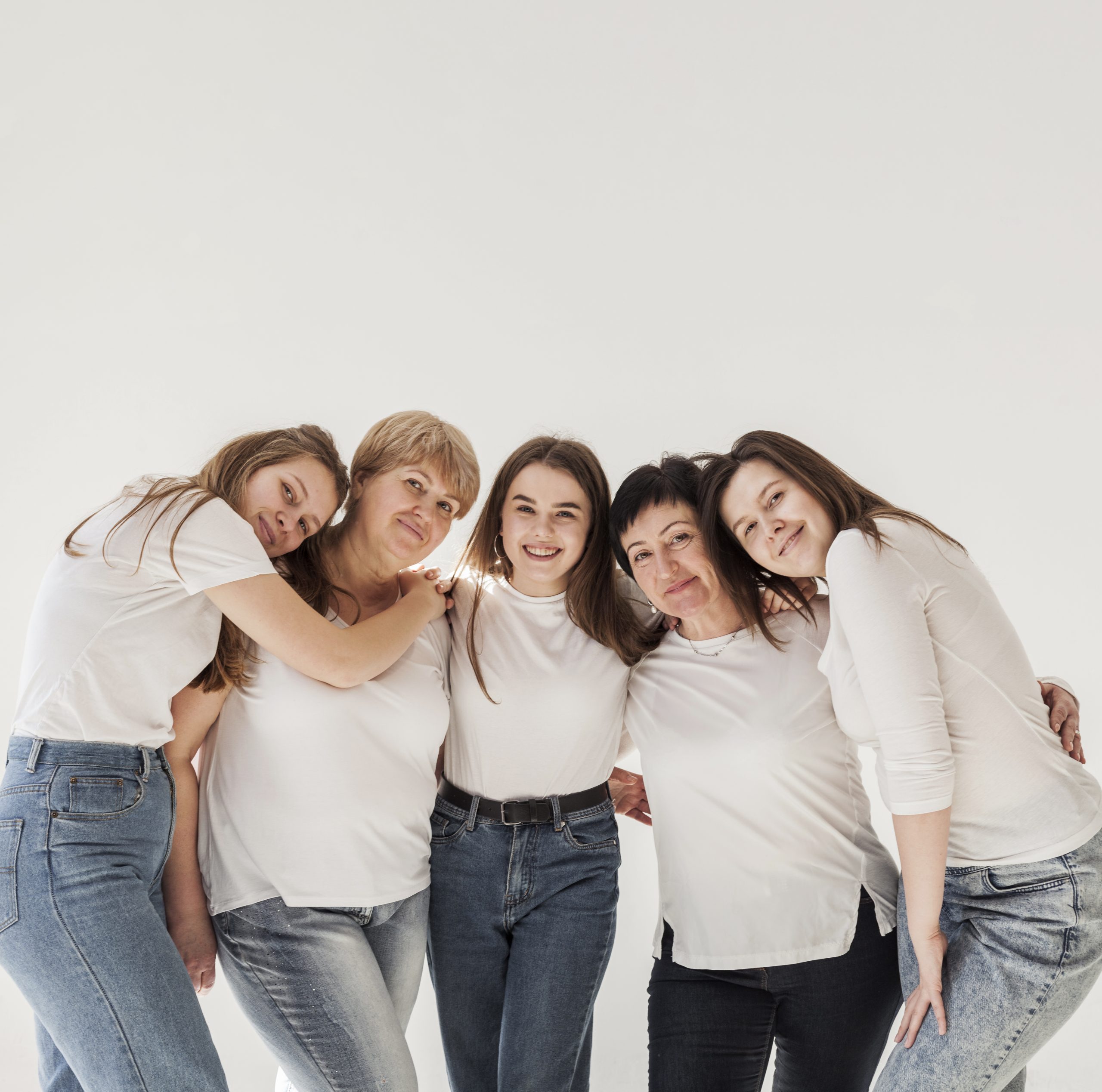 Fünf Frauen unterschiedlicher Altersgruppen umarmen sich vor weißem Hintergrund. Sie tragen weiße T-Shirts und Jeans.